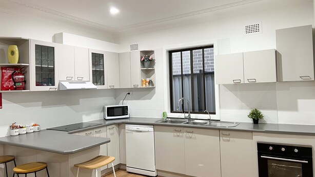 Una cocina moderna con armarios blancos, un microondas integrado, un lavavajillas y una estufa. Hay dos taburetes y una ventana con persianas.