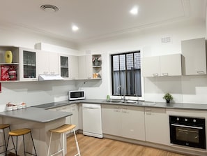 A modern kitchen with white cabinetry, a built-in microwave, a dishwasher, and a stove. There are two stools and a window with blinds. - This one storey house has 5 bedrooms and 2 full bathrooms (Fairfield Heights)