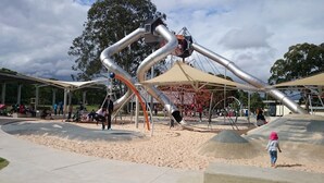 Un parque infantil con una gran estructura para escalar, zona de juegos con arena y asientos cubiertos.