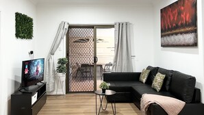 Una sala de estar moderna con un sofá negro, un televisor sobre un mueble bajo, una mesita y una planta.
