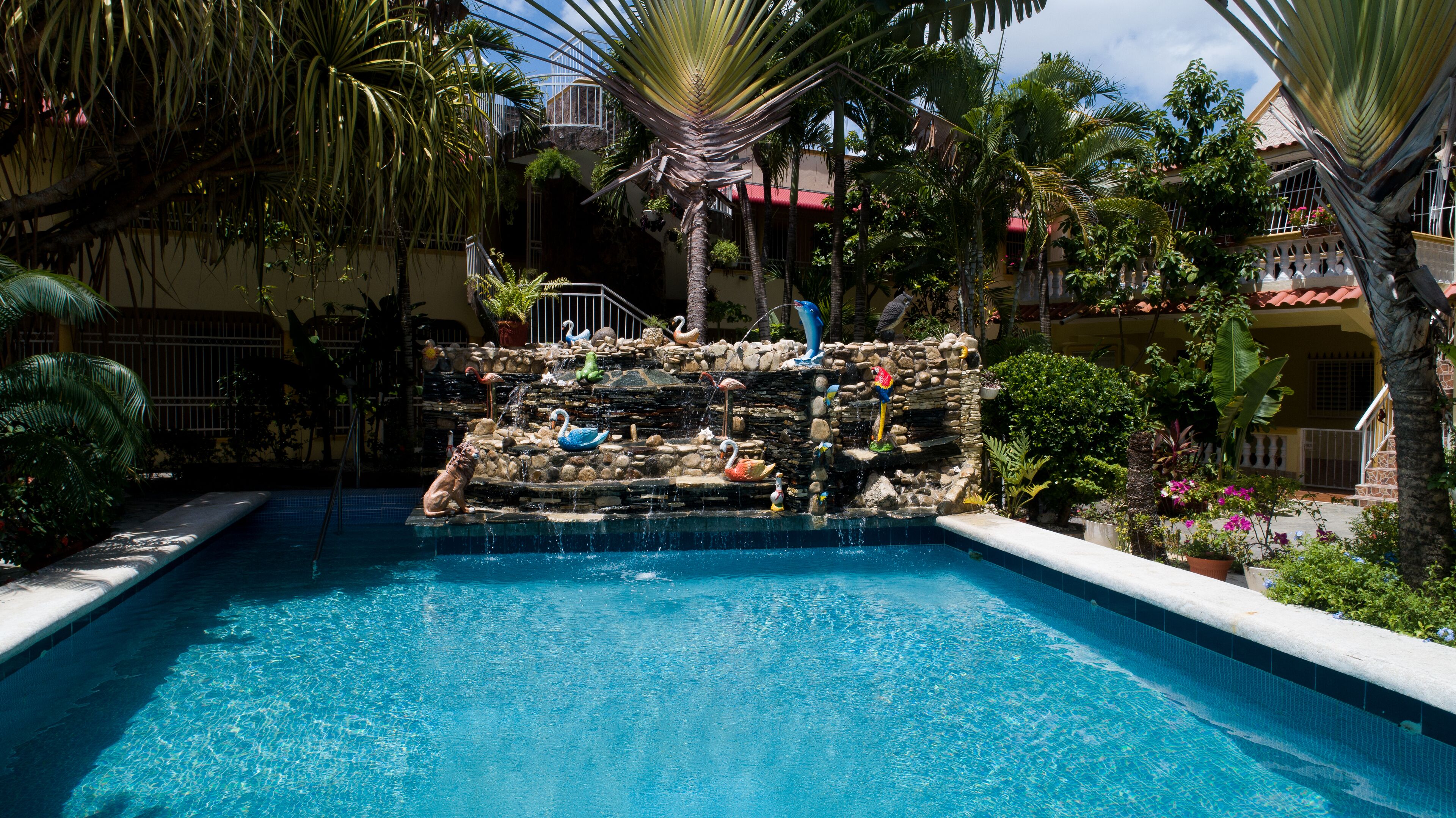 A swimming pool with a waterfall feature, surrounded by tropical plants and a stone wall with decorative swans.