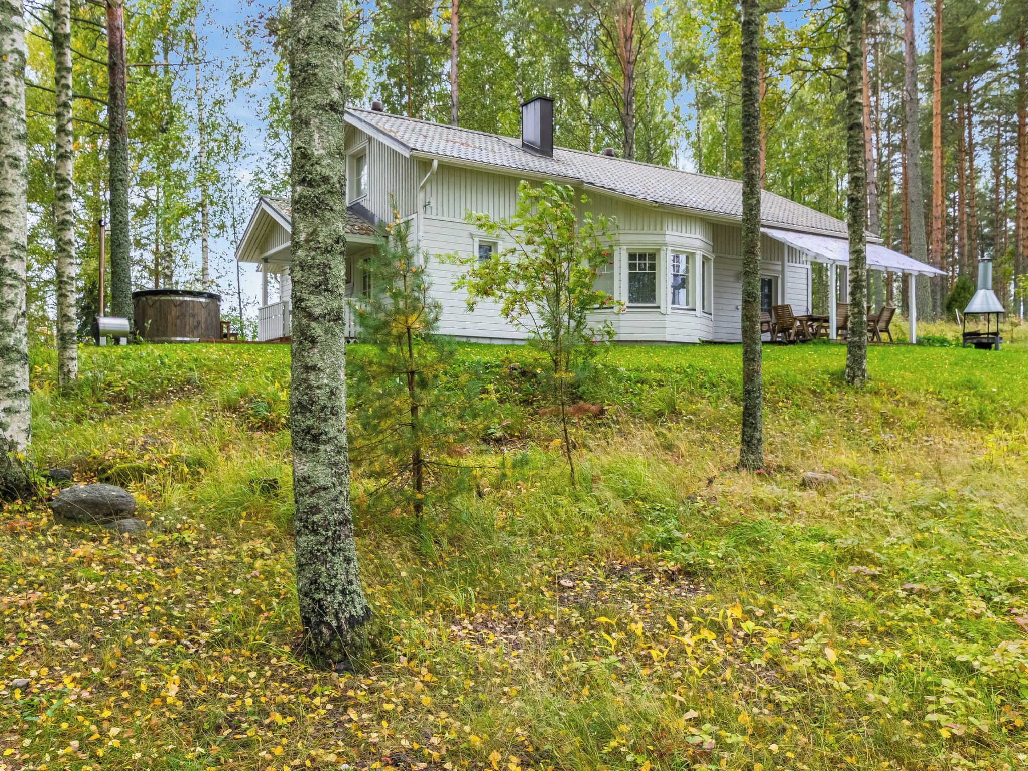 A white cabin with a covered porch, surrounded by a forested area with trees and a grassy field.