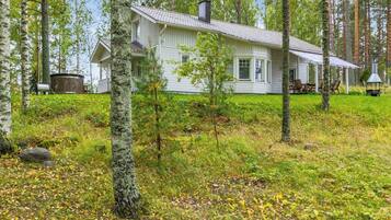 A white cabin with a covered porch, surrounded by a forested area with trees and a grassy field.