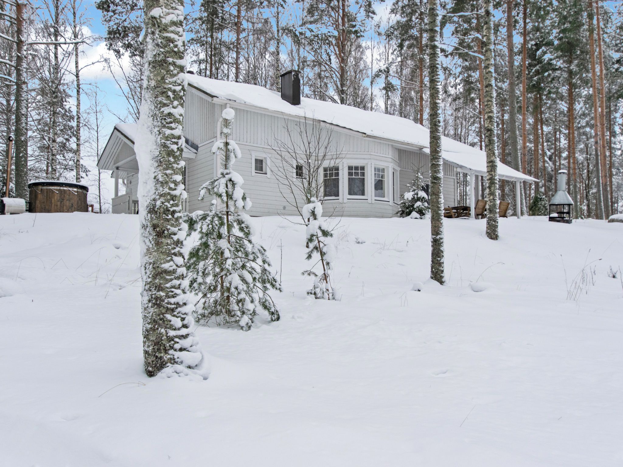A snow-covered wooden house with a garage, surrounded by tall pine trees.