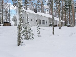 A snow-covered wooden house with a garage, surrounded by tall pine trees.
