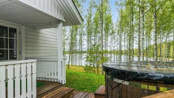 A wooden deck with a hot tub, a porch with a white railing, and a view of a lake with birch trees.