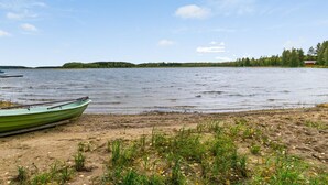 Escena junto al lago con un bote verde en la orilla, una playa de arena y un área boscosa al fondo.
