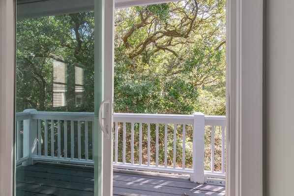 A view through a glass door onto a wooden deck with white railings, surrounded by lush greenery.