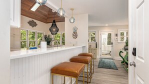 Une cuisine avec des meubles blancs, un plafond en bois et des luminaires suspendus. On y trouve trois tabourets et un banc vert.