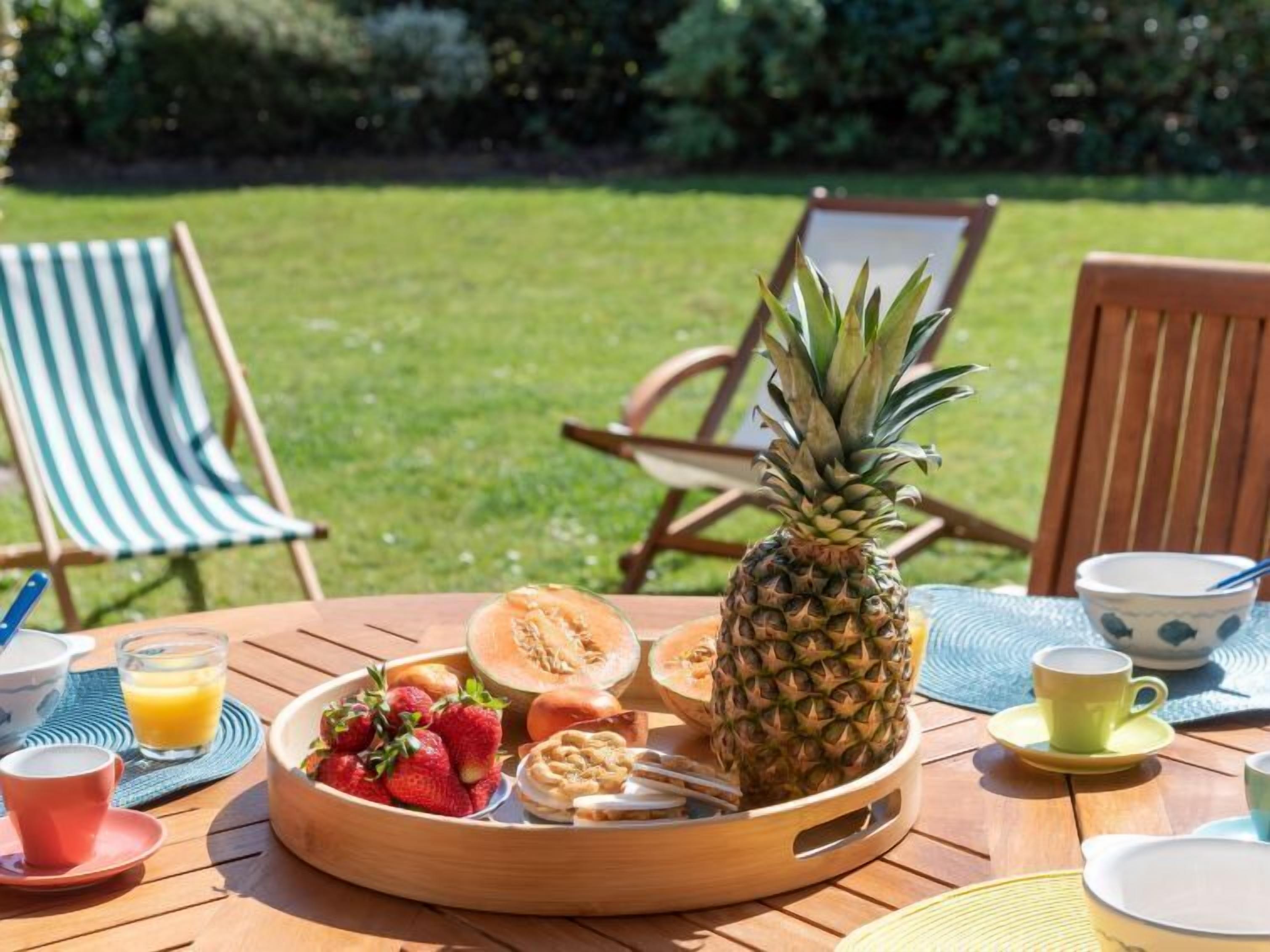 Uma mesa de madeira com um prato de frutas, uma abacaxi e um copo de suco de laranja.