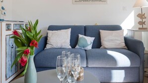 Sala de estar com um sofá azul, almofadas decorativas, um vaso com flores e uma mesa com copos.