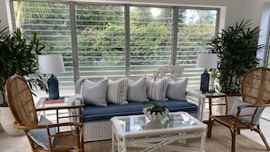 A cozy living room with a blue sofa, wicker chairs, a glass coffee table, and large windows overlooking greenery.