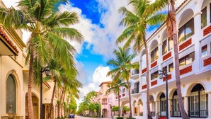 A street lined with palm trees and buildings with balconies and arched windows.