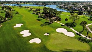 A golf course with sand bunkers, a water body, and residential areas in the background.