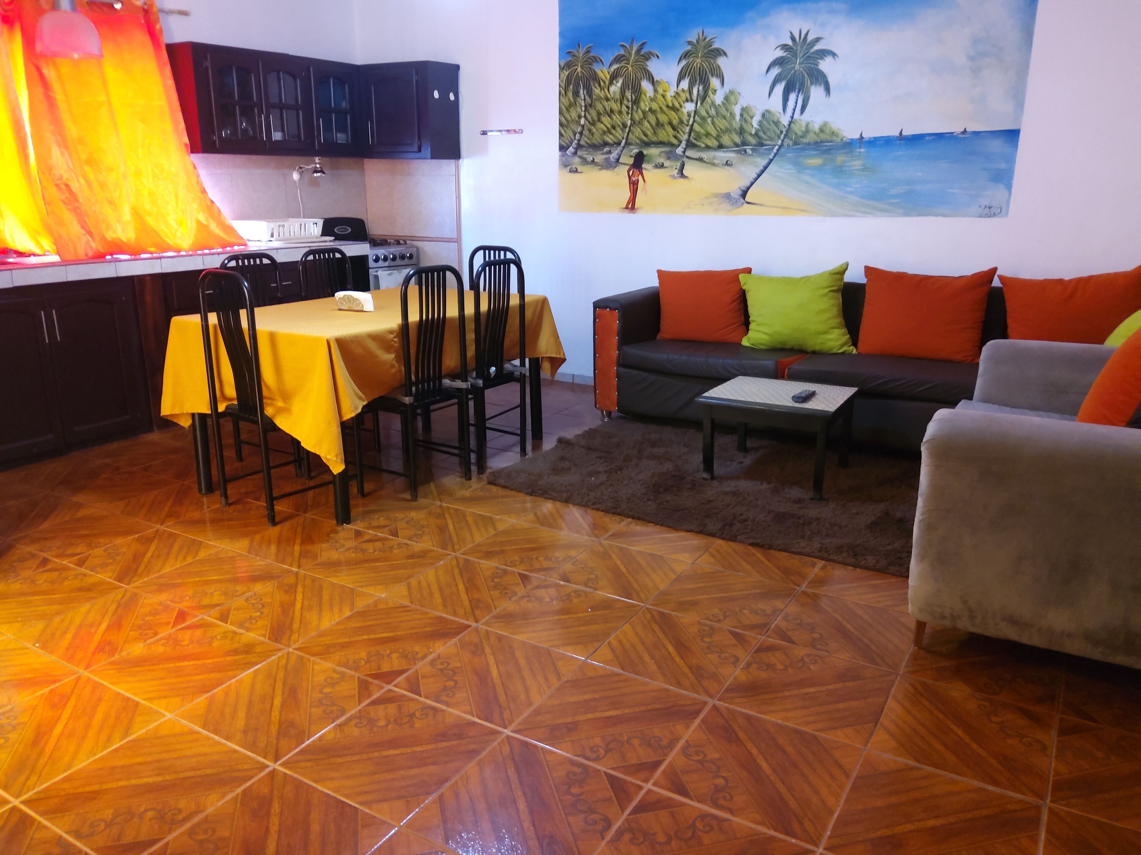 Un área de comedor con una mesa y sillas, un sofá con cojines, un cuadro de una playa y una cocina con armarios y estufa.