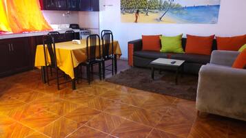 Un área de comedor con una mesa y sillas, un sofá con cojines, un cuadro de una playa y una cocina con armarios y estufa.