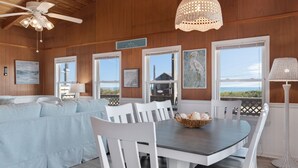 A dining area with a round table, white chairs, and a chandelier. There is a view of the beach through large windows. - SEMI OCEANFRONT, Steps to the Beach, Unobstructed Amazing Ocean Views. Remodeled (Kitty Hawk)