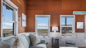 A cozy room with a wooden ceiling, a grey sofa with patterned cushions, wicker chairs, and a view of the beach. - SEMI OCEANFRONT, Steps to the Beach, Unobstructed Amazing Ocean Views. Remodeled (Kitty Hawk)