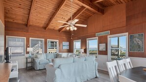 A spacious living room with a high ceiling and wooden beams, featuring a light blue sofa set, a dining table with chairs, and a ceiling fan. - SEMI OCEANFRONT, Steps to the Beach, Unobstructed Amazing Ocean Views. Remodeled (Kitty Hawk)