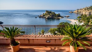 A balcony with potted palm plants, overlooking a coastal view with rocky outcrops and a beach.
