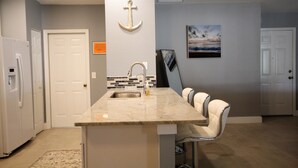 A modern kitchen with a marble countertop, a sink, and a bar area with two bar stools. There is a white refrigerator on the left and a door with a white handle. - Newly Renovated, Beautiful Spacious House With Heated Pool. (Englewood)