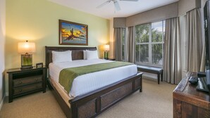 A hotel room with a large bed, a ceiling fan, a painting on the wall, and a window with curtains.