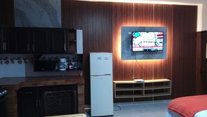 Une chambre d’hôtel compacte comprenant un coin cuisine, un téléviseur à écran plat fixé au mur, un lit avec une couverture rouge et un petit coin salon.