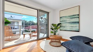 A modern living room with a large sliding glass door leading to a balcony, wicker furniture, a round woven table, and a large abstract painting on the wall. - Coast Bliss on Currambene (Huskisson)
