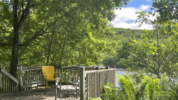 Une terrasse en bois, agrémentée de bancs et d’une chaise jaune, offre une vue sur une rivière et est entourée d’une végétation luxuriante.