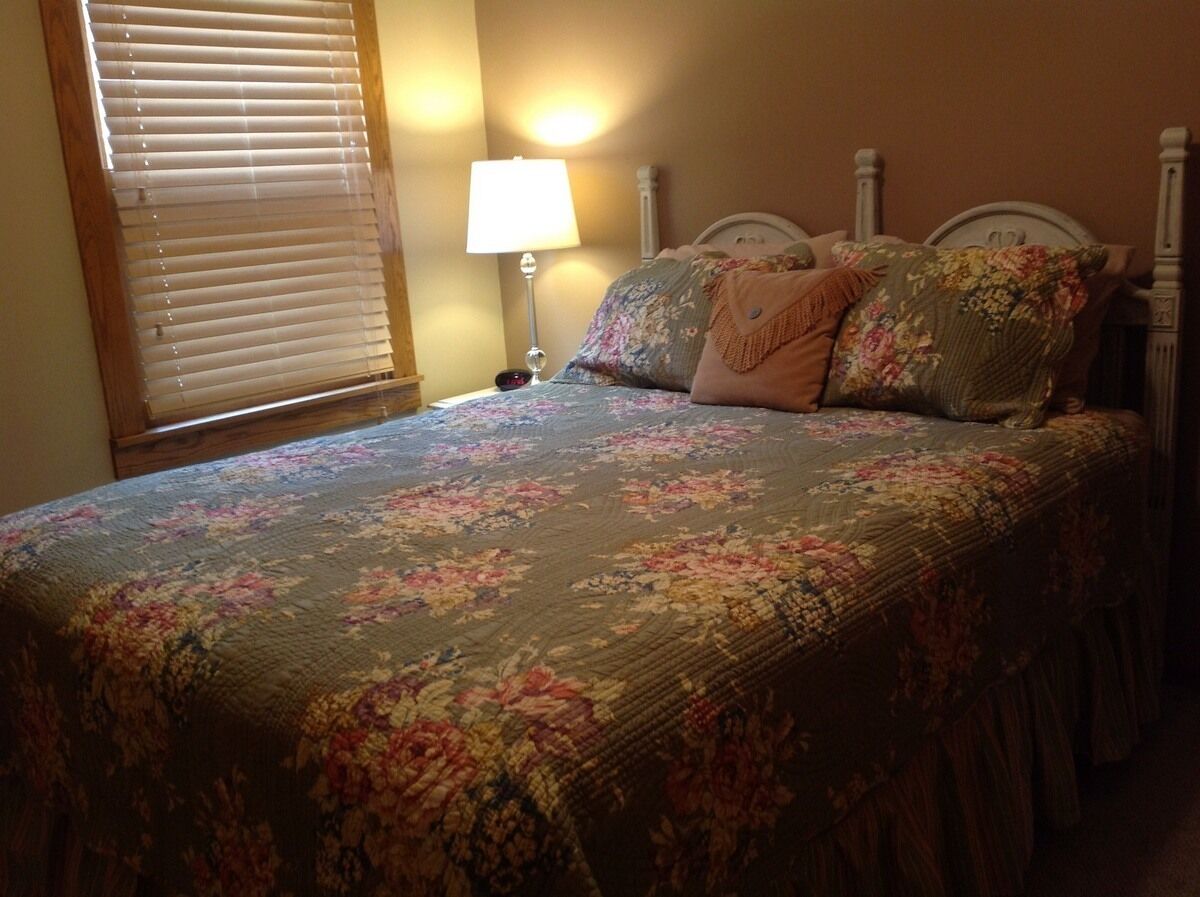 Un lit avec une couvre-lit à motifs floraux, une lampe de chevet et une fenêtre avec des stores.