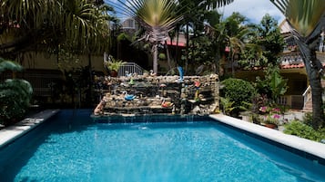 Una piscina con cascada, rodeada de plantas tropicales y un muro de piedra con cisnes decorativos.