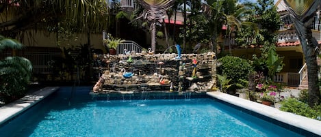 A swimming pool with a waterfall feature, surrounded by tropical plants and a stone wall with decorative swans.