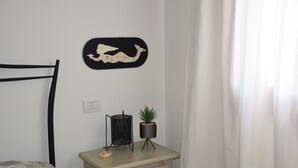A bedroom with a bed, a nightstand with a plant, a wall-mounted artwork, and a curtain. - The House in Sardinia Holiday Apartment (Olbia)