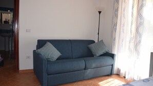 A room with a dark blue sofa, patterned cushions, a standing floor lamp, and a window with sheer curtains. - The House in Sardinia Holiday Apartment (Olbia)