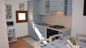 A compact kitchen with light blue cabinets, a gas stove, and a microwave. There is a window with a wooden frame and a small shelf with decorative items. - The House in Sardinia Holiday Apartment (Olbia)