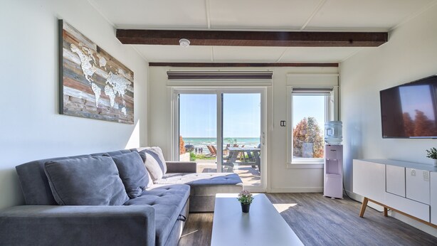 Ein modernes Wohnzimmer mit grauem Sofa, einem weißen Couchtisch und einer Weltkarte als Wandbild. Es gibt ein großes Fenster mit Blick auf den Strand und einen Wasserspender.