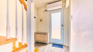 Ein schmaler Flur mit einer Holztreppe, einer Tür mit blauer Fußmatte und einer Klimaanlage an der Wand.