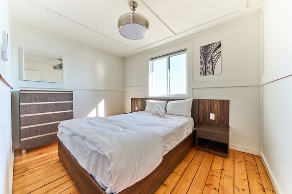 Une chambre avec un plancher en bois, un lit recouvert de linge de lit blanc, une table de chevet, un miroir fixé au mur et une fenêtre donnant sur des arbres.