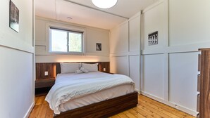 Ein Schlafzimmer mit Holzboden, einem Bett mit weißer Bettwäsche, einem Nachttisch, einem Fenster und einer Deckenleuchte.