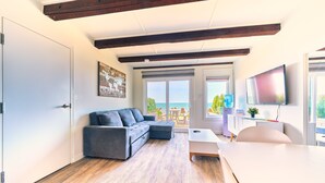 Ein modernes Wohnzimmer mit einem grauen Ecksofa, Holzböden, einem Flachbildfernseher und einem großen Fenster mit Meerblick.