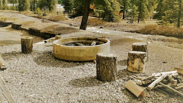 A campsite with a fire pit surrounded by wooden stumps and logs, set in a forested area with mountains in the background.