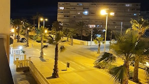 A nighttime view of a street with a white car parked on the side, palm trees, and a building with illuminated windows in the background. - Sea view holiday apartment (Torrevieja)