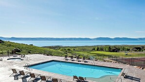 A swimming pool with lounge chairs and a tennis court, surrounded by a fence and a view of a lake and mountains. - Private beach, pools/hot tub, winter sports, sleeps 20 (Fish Haven)