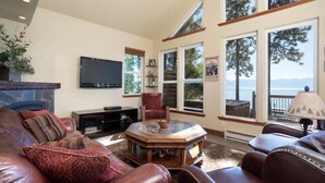 A spacious living room with leather sofas, a fireplace, a flat-screen TV, and large windows offering a view of the outdoors.