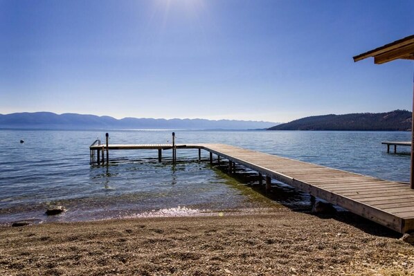 A wooden pier extending into calm water with clear skies and distant mountains.