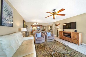 A living room with a sofa, armchairs, a glass coffee table, a ceiling fan, a TV, and a wooden cabinet.