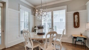 A dining area with a round table, white chairs, a chandelier, and a view through large windows. - Luxury 4BR Home! Walk to Beach & Bike to Pool! ~ The Wander Inn at WaterSound (Santa Rosa Beach)
