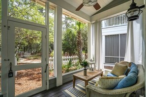 A covered porch with a view of a garden, a wicker chair, a table, and a ceiling fan.