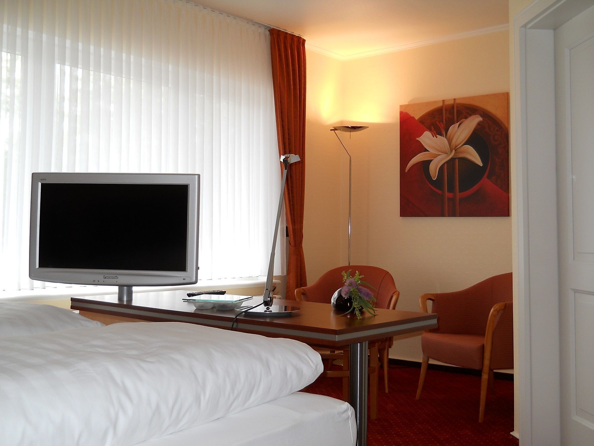 A hotel room with a bed, a television, a desk with a lamp, a chair, and a painting on the wall.