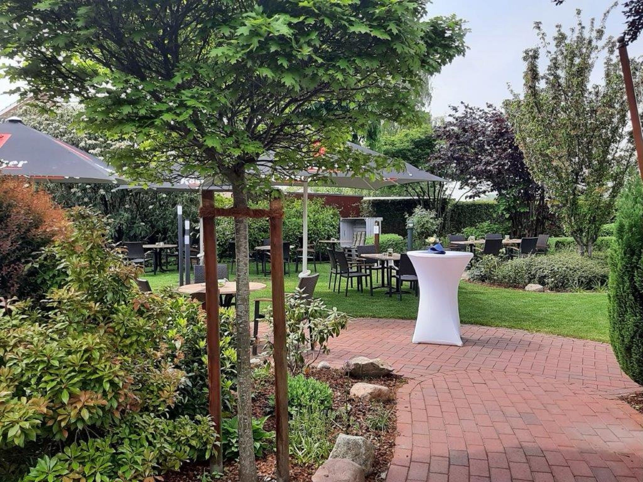 Eine Terrassenfläche im Freien mit Tischen und Stühlen, umgeben von Pflanzen und Bäumen.